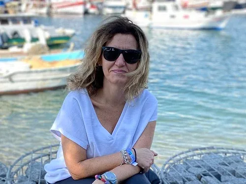 Aldara Laso, con gafas de sol, sentada junto al agua de un puerto con barcas de pescadores