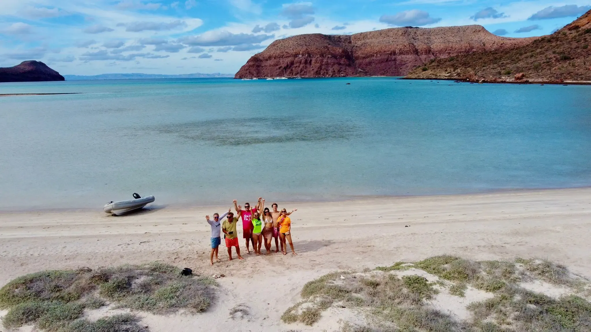 Grupo de la flotilla saludando en una playa de arena blanca, con la neumática en la orilla y acantilados rojizos al fondo