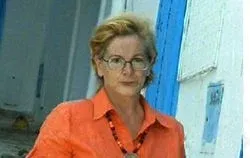 Maite Pérez, con gafas y blusa naranja, junto a una casa encalada de puertas azules