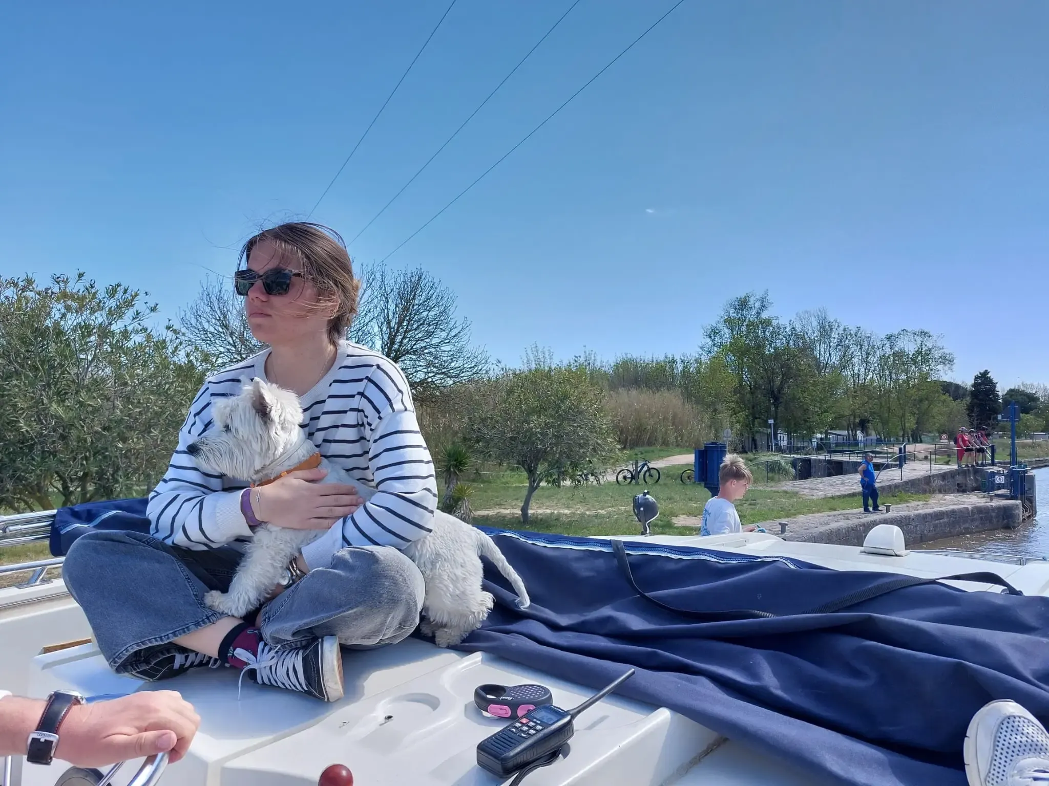 Navegante con gafas de sol y un perro blanco en brazos, sentada en la cubierta de proa del barco, con una esclusa al fondo