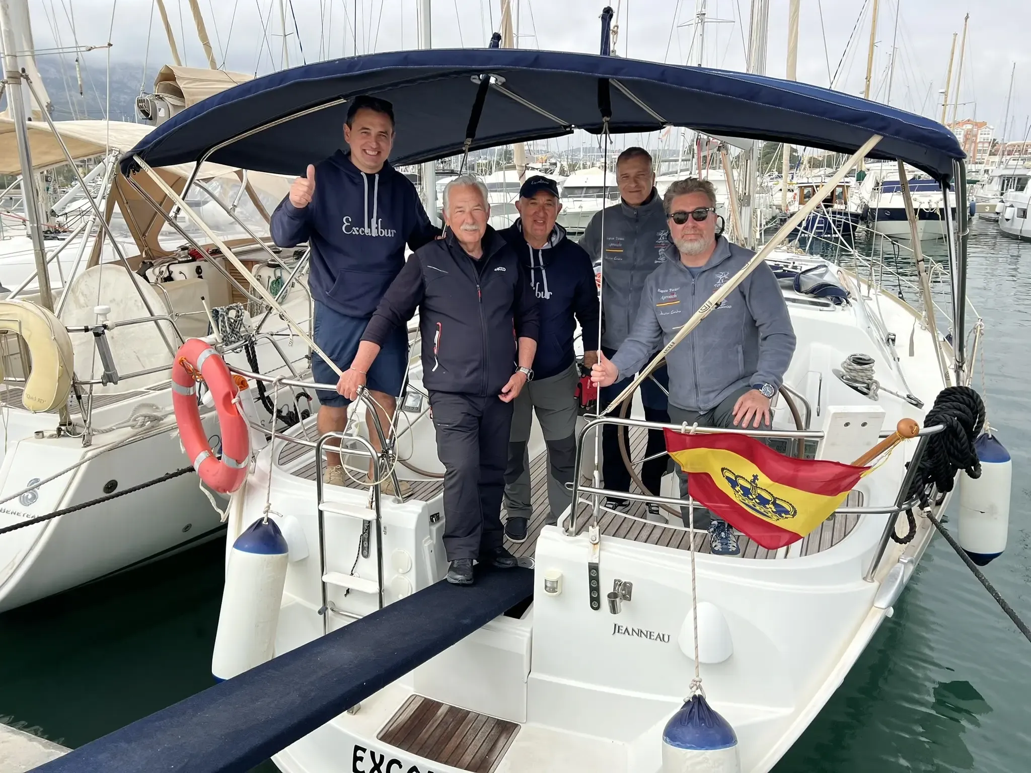Tripulación posando en la proa de un velero amarrado en el puerto, con una bandera de España