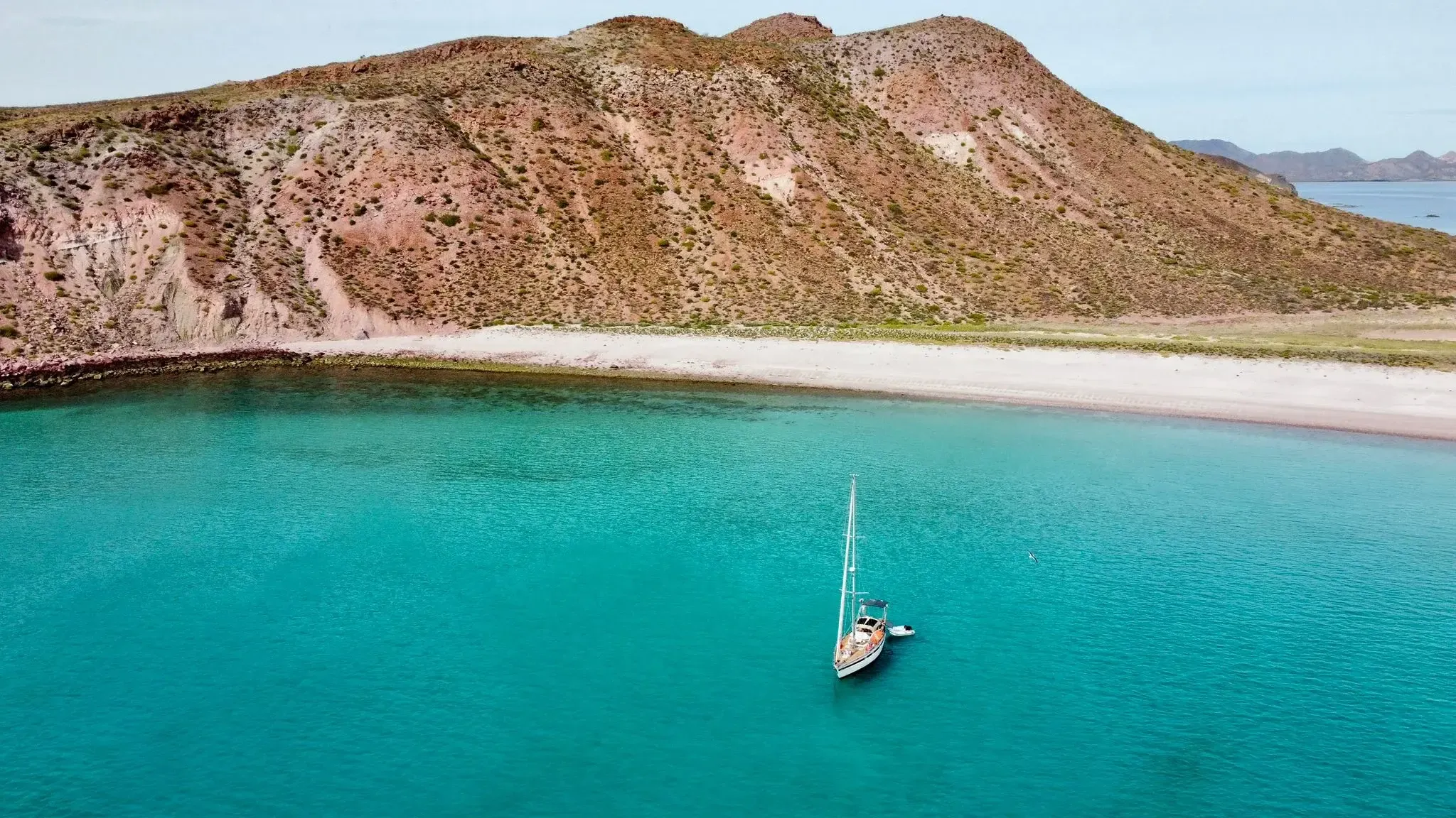 Velero fondeado en aguas turquesa al pie de una montaña rojiza, junto a una playa de arena blanca